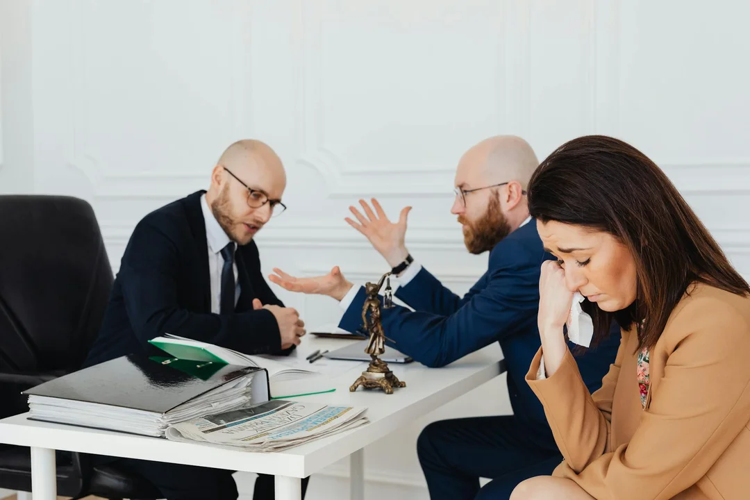 Three professionals sit at a white office desk discussing legal documents, with a small statue of Lady Justice on the table.