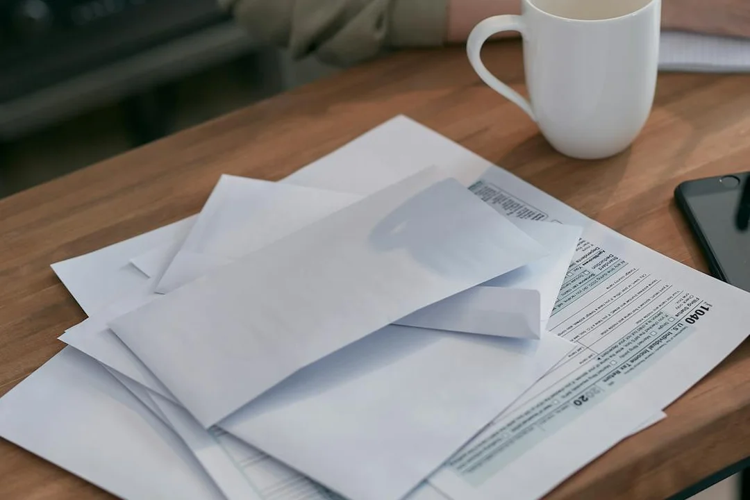 Desk with stacked envelopes and documents, a coffee mug, and a smartphone, suggesting administrative paperwork and financial planning