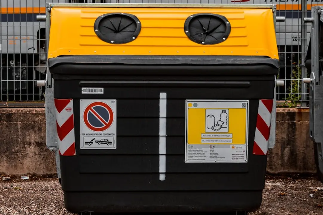 A large black and yellow dumpster with warning labels sits in front of a fence.