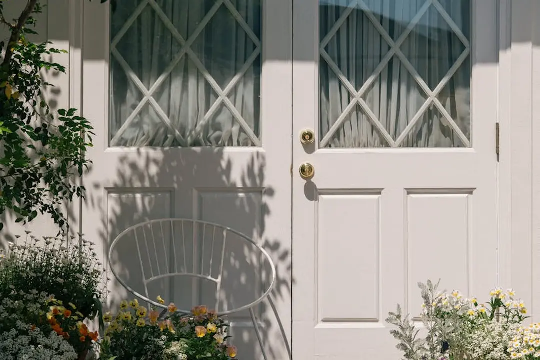 White front door with diamond-pattern glass panes, a metal chair, and surrounding plants