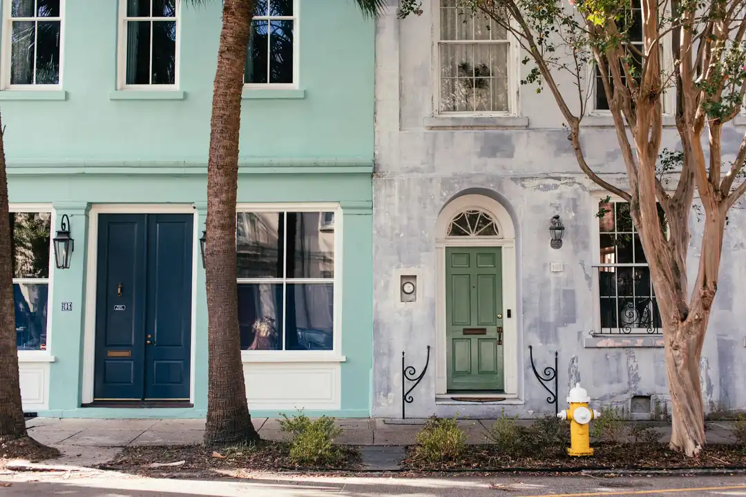 Row of pastel townhouses with green doors, trees along the sidewalk, and a yellow fire hydrant in front.