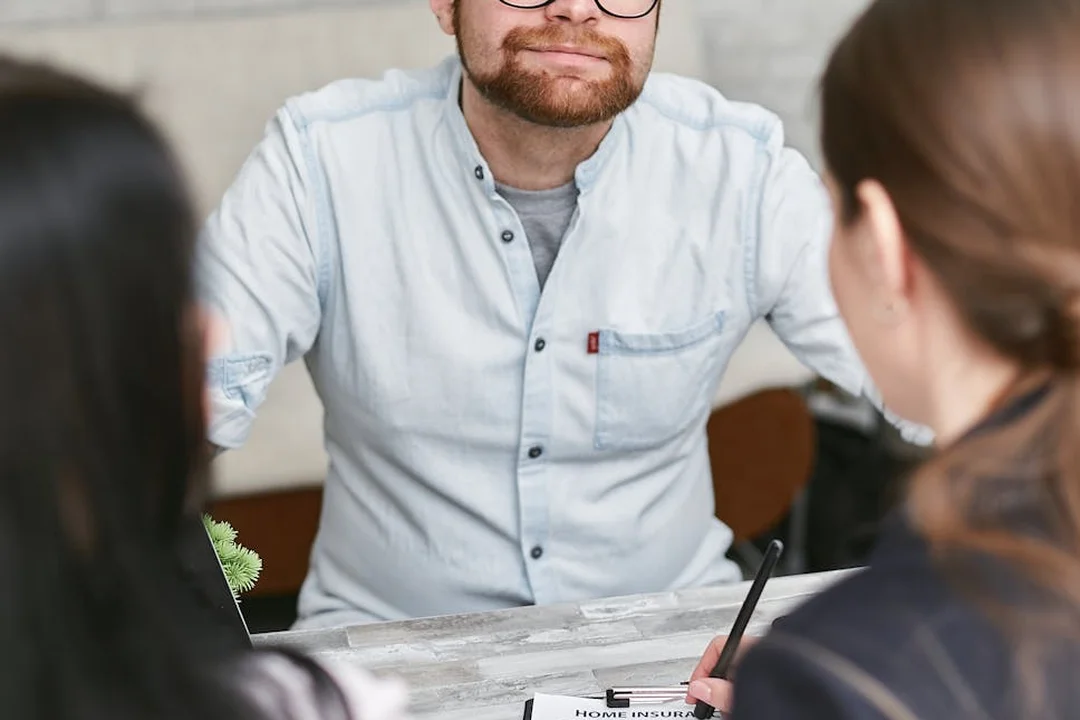 A professional consultant wearing a light blue shirt and glasses discusses documents with two clients across a table.