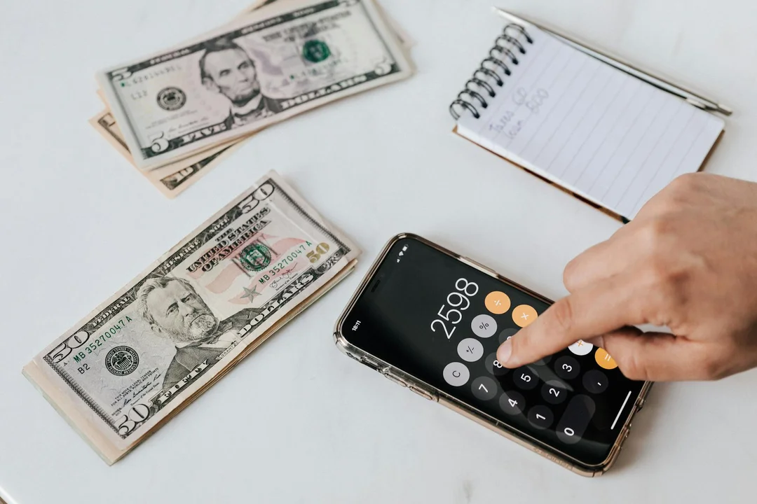 A hand using a calculator on a desk with US dollar bills and a notebook, symbolizing cost verification for HOA fees on home listings.