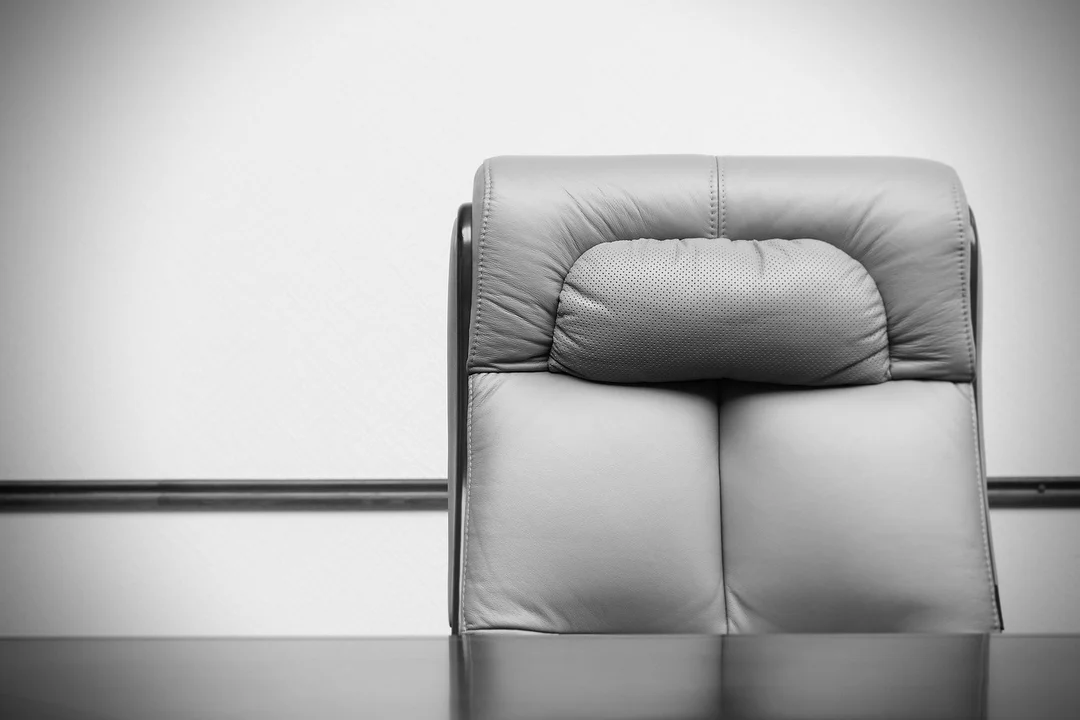 Black-and-white image of a padded office chair backrest above a dark desk, in a minimalist office setting