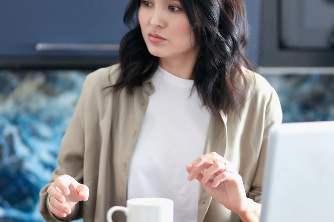 Woman in a beige blazer sits at a desk with a laptop and a coffee mug, looking thoughtful while discussing figures.