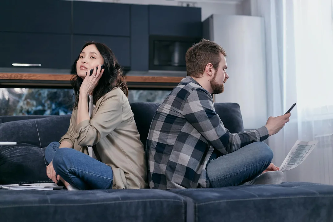 A couple sits on a modern living room sofa; the woman on the left is talking on her phone and looking away, while the man on the right holds a sheet of paper and a phone, conveying financial tension.
