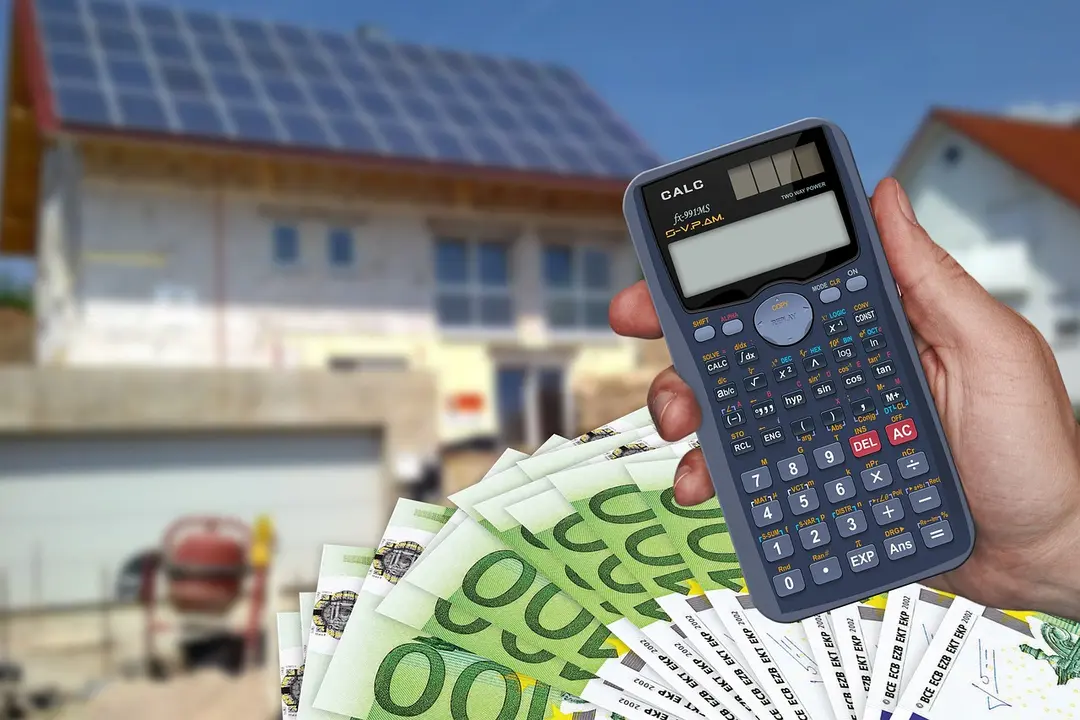 A hand holds a calculator in the foreground with a fan of euro banknotes, and a suburban house with solar panels in the blurred background.