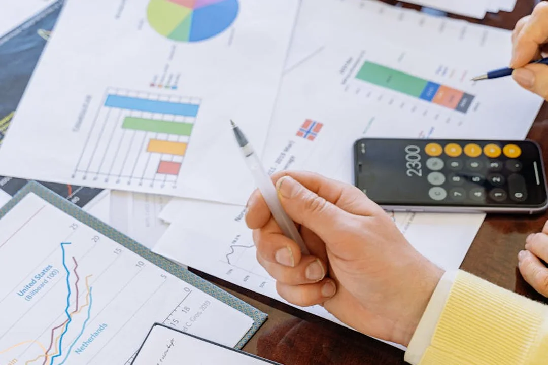A close-up of charts and graphs on a desk, with a hand holding a pen and a calculator nearby, illustrating financial analysis of HOA fees and mortgage impact.