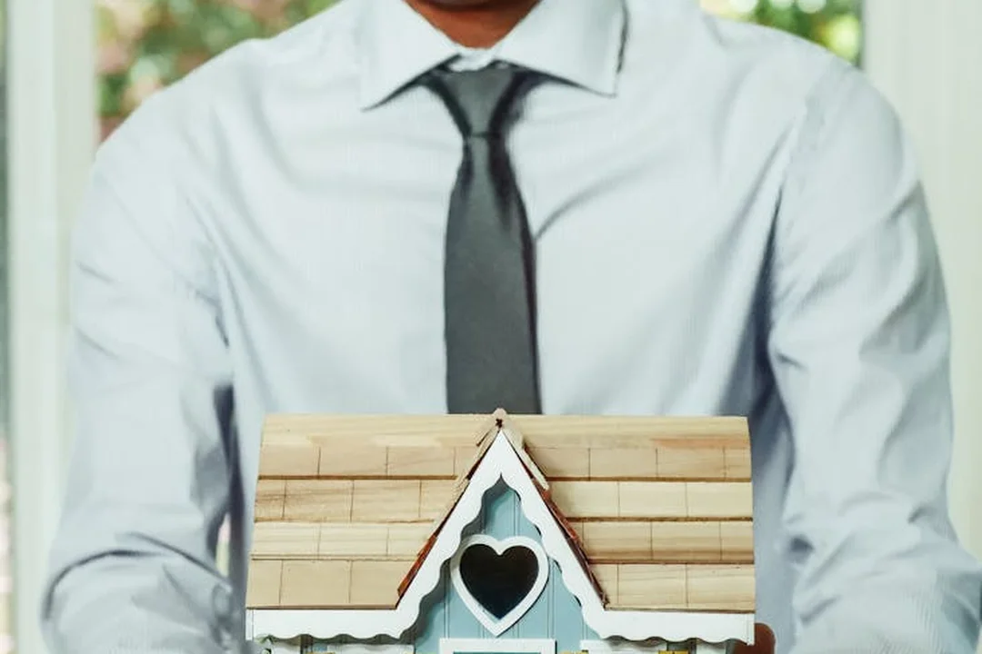 A person in a light blue shirt and dark tie holds a small house model with a heart-shaped window.