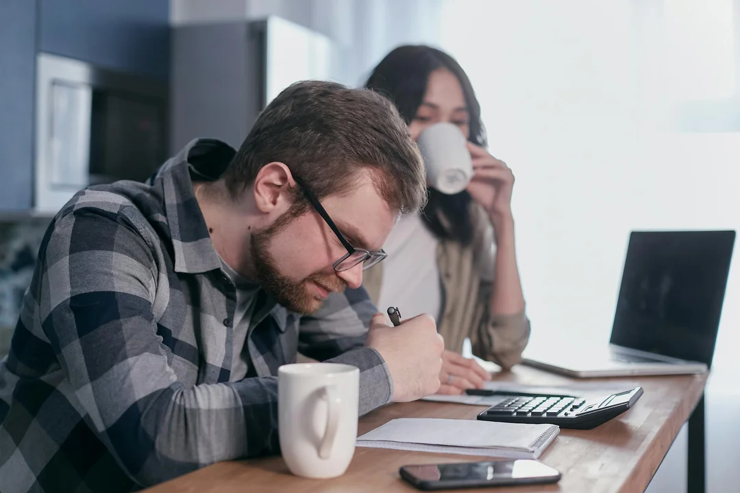 Two people sit at a wooden table reviewing documents. One writes on papers with a pen while the other sips from a mug; a calculator, smartphone, and laptop are on the table.