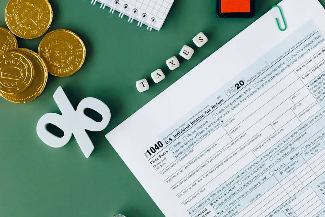 Close-up of tax-related items: gold coins, a white decorative tax symbol, a notebook, and a form with printed lines, with letter blocks spelling TAXES in the background, representing HOA tax filing concepts.