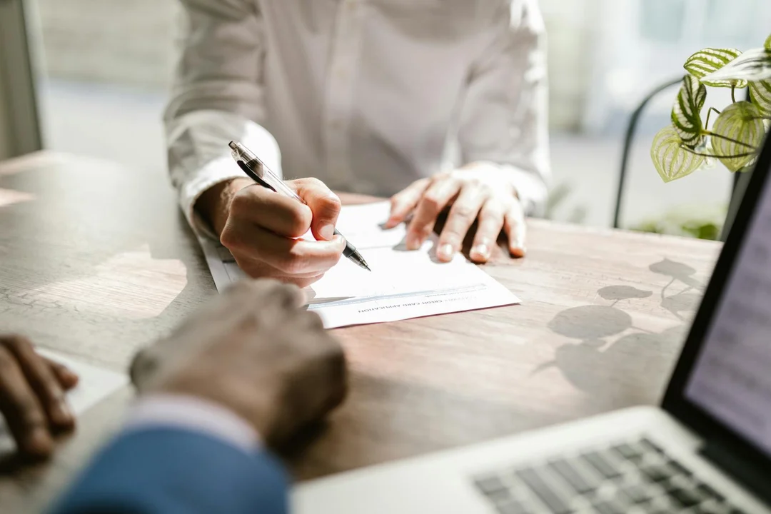 Close-up of hands signing a document at a desk, with a laptop and a plant in the background.