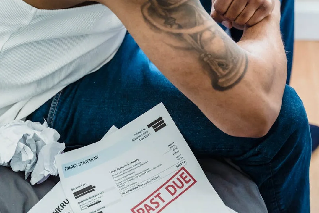 A person with a tattooed arm sits on a couch reviewing past-due bills with a red 'PAST DUE' stamp on the papers.