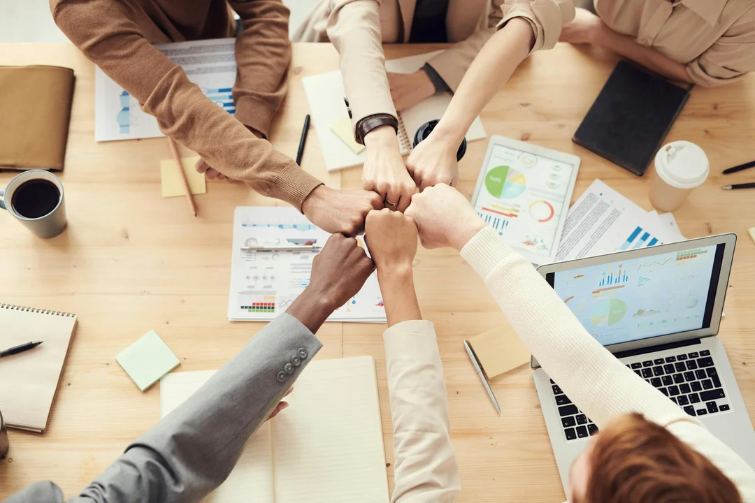 Group of people joining hands in a circle over a table with documents, charts, and a laptop.