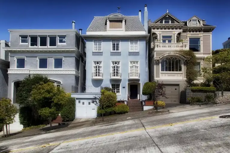 Row of pastel-colored townhouses on a steep urban street with garages and greenery.