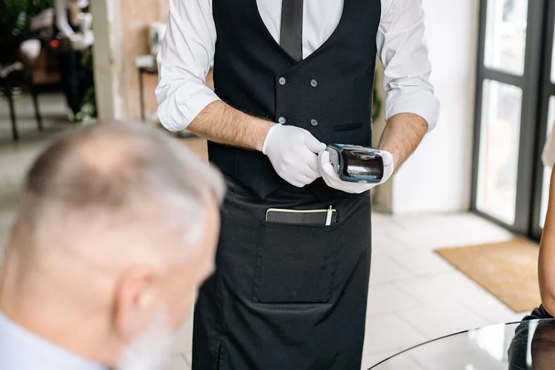 A staff member in a black vest and white shirt uses a card reader to process a payment for a customer at a table.