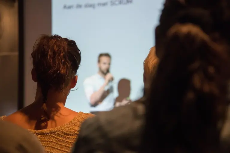 A speaker on stage delivering a presentation to an audience, with attendees shown from behind in a dimly lit room.