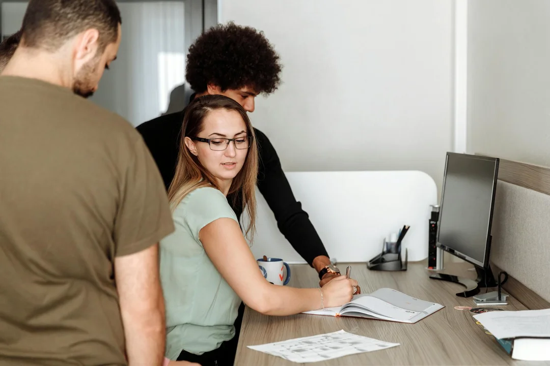 Three people in an office around a desk with documents and a computer monitor, discussing and reviewing materials