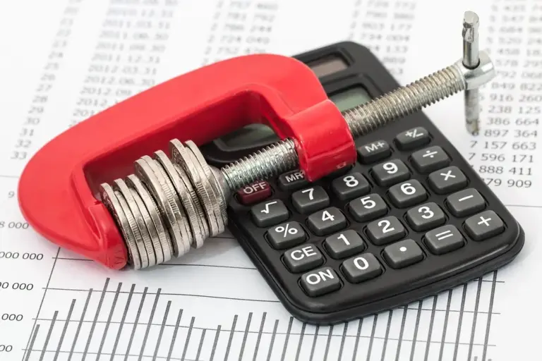 A red C-clamp grips a stack of coins resting on a calculator, with financial charts in the background, illustrating the financial and legal aspects of HOA reserve funds.