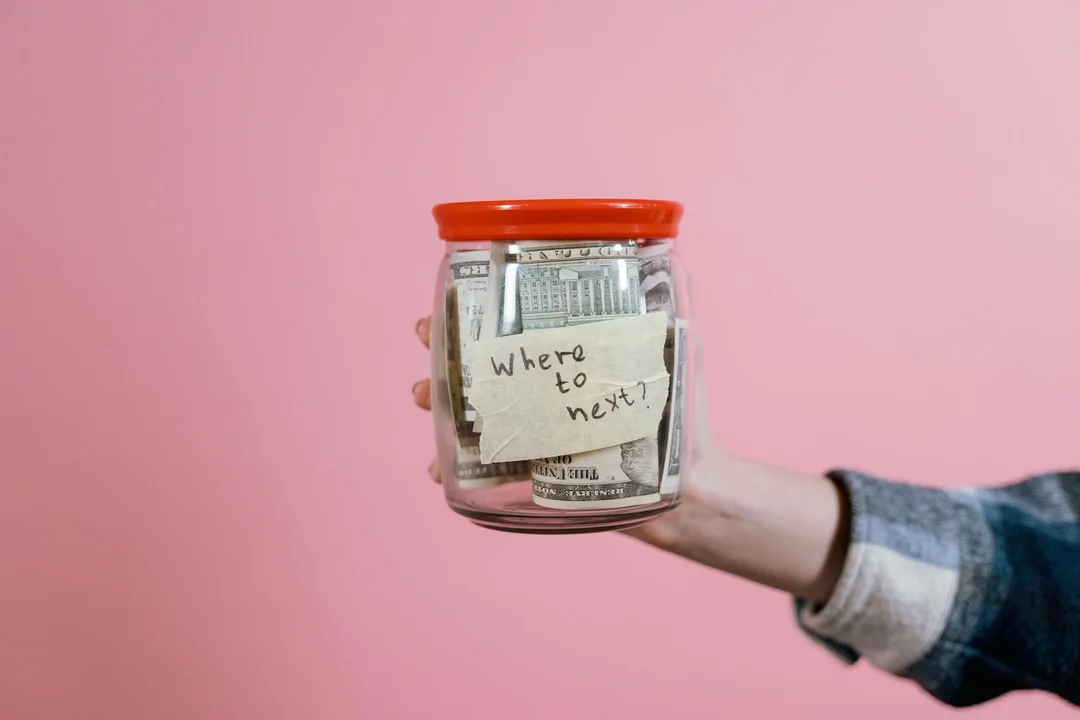 Hand holding a glass jar filled with money against a pink background, with a sticky note reading 'Where to next?'