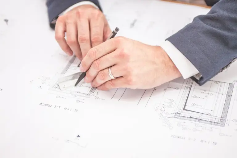 Close-up of hands in a suit drawing on architectural blueprints with a pencil and scale.