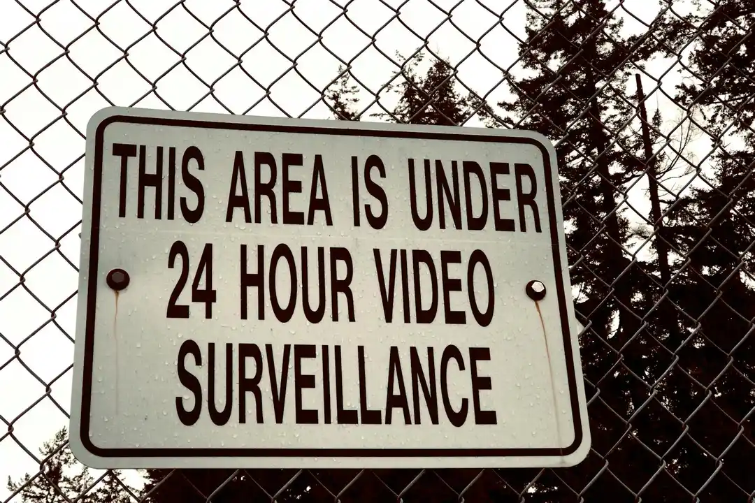 Sign stating 'This area is under 24 hour video surveillance' mounted on a chain-link fence with trees in the background.