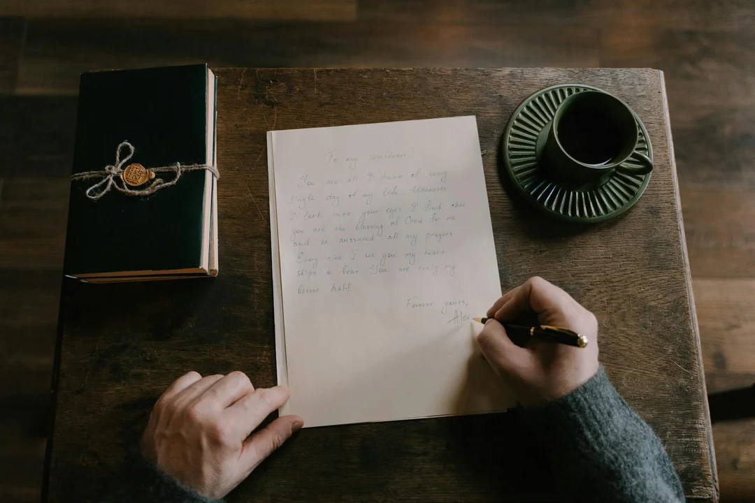 Top-down view of a person writing on a sheet of paper at a wooden desk, with a closed book tied with string on the left and a dark cup on a saucer on the right.