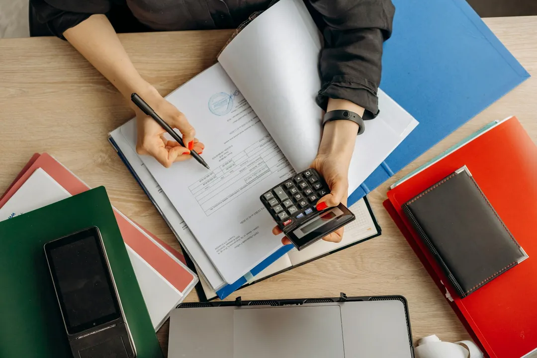 Overhead view of a person at a wooden desk reviewing documents, writing with a pen, and using a calculator, surrounded by colorful folders and notebooks—an illustration of HOA amendment work.