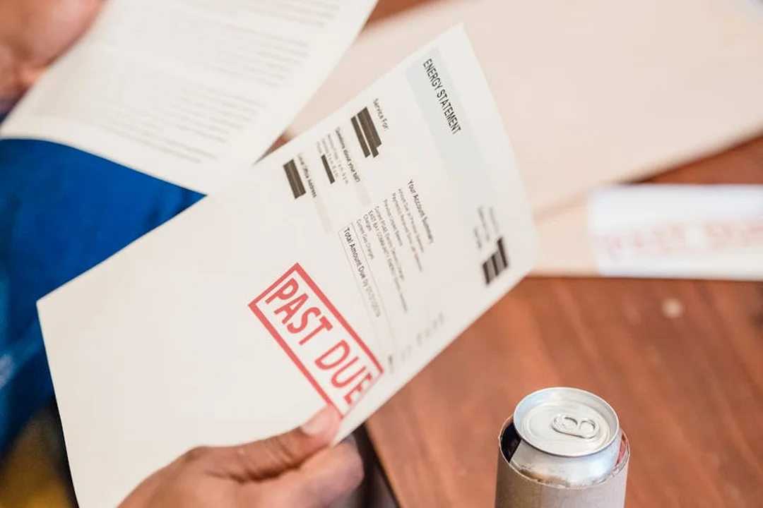 Person's hand holding a document stamped 'PAST DUE' beside a can on a wooden table.