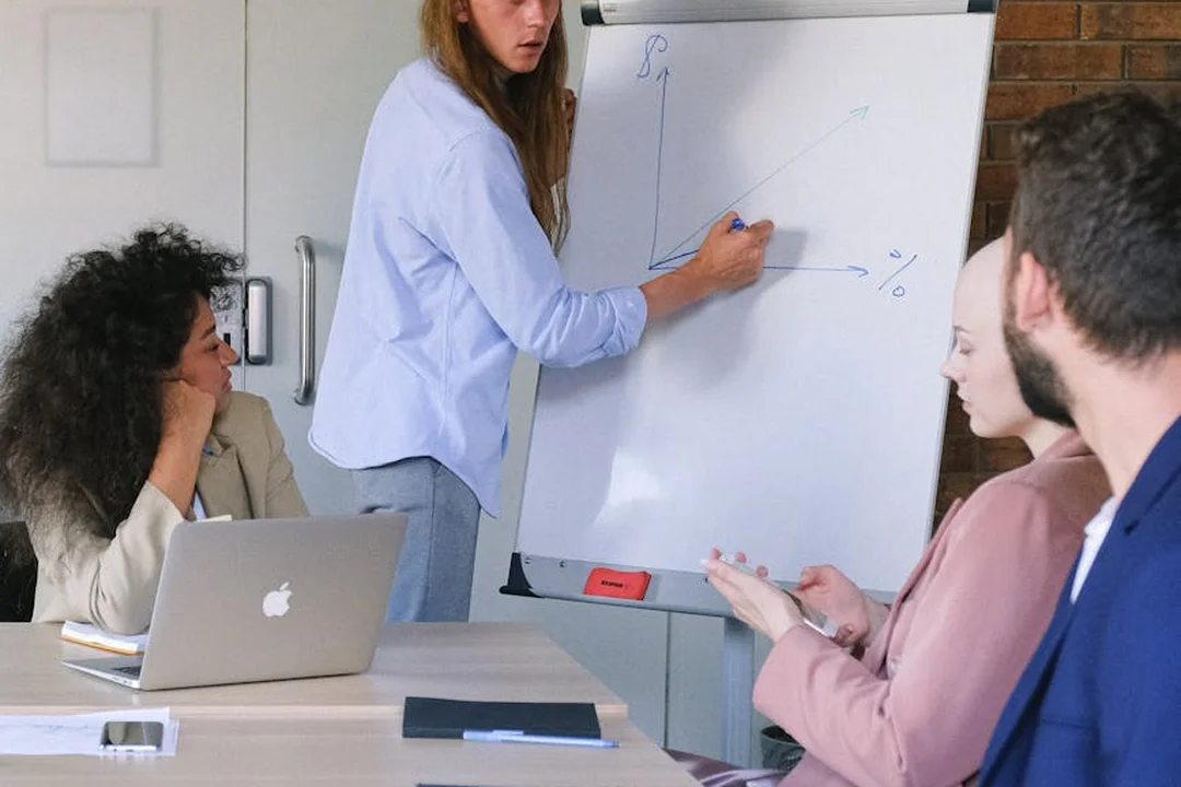 Group of professionals in a meeting around a table; a presenter writes on a whiteboard as others listen and take notes.