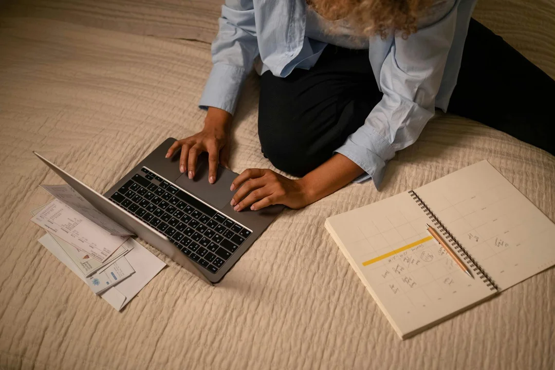 Person wearing a light blue shirt sits on a carpeted floor, typing on a laptop with a notebook and scattered documents beside them, representing planning during the first 90 days on an HOA board.
