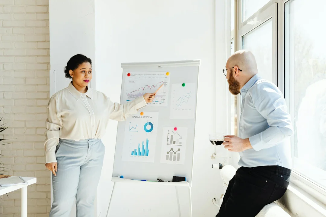 A woman presents charts on a flip chart while a man drinks coffee and listens in a bright office, illustrating governance discussion.