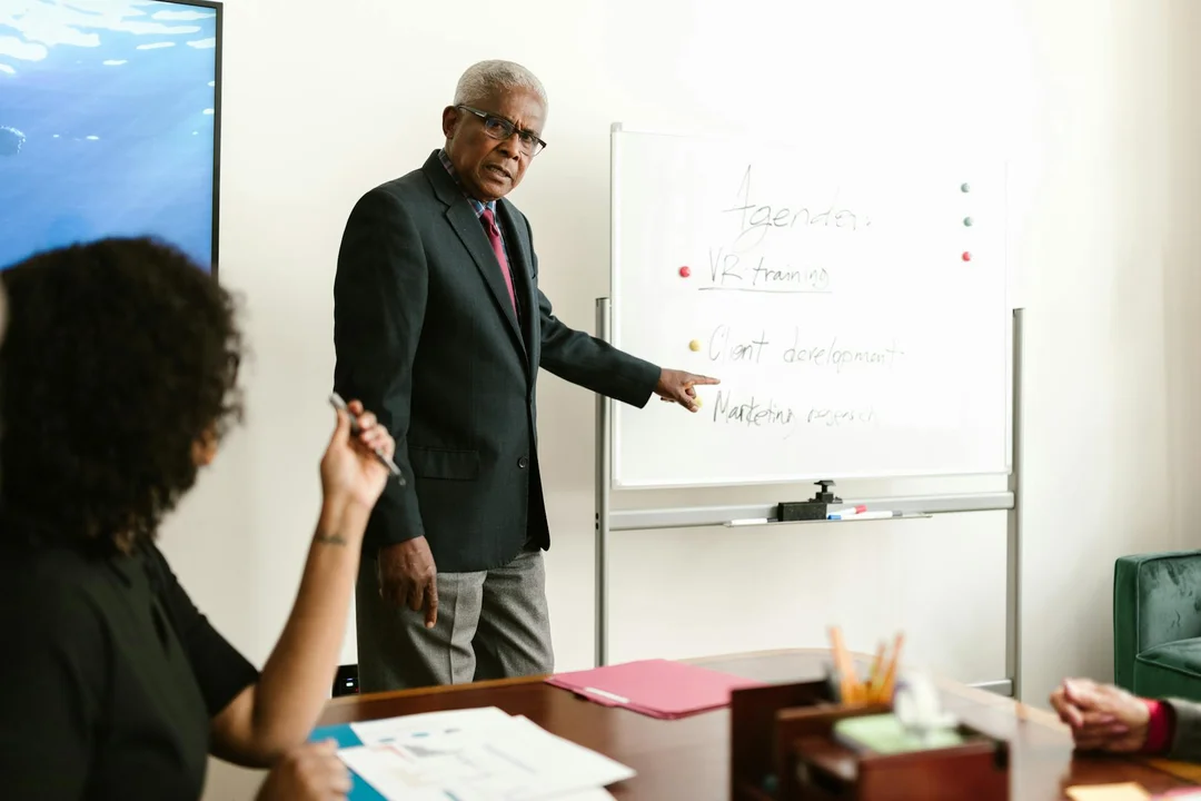 A presenter in a dark suit points to a whiteboard during a boardroom training session about HOA governance.