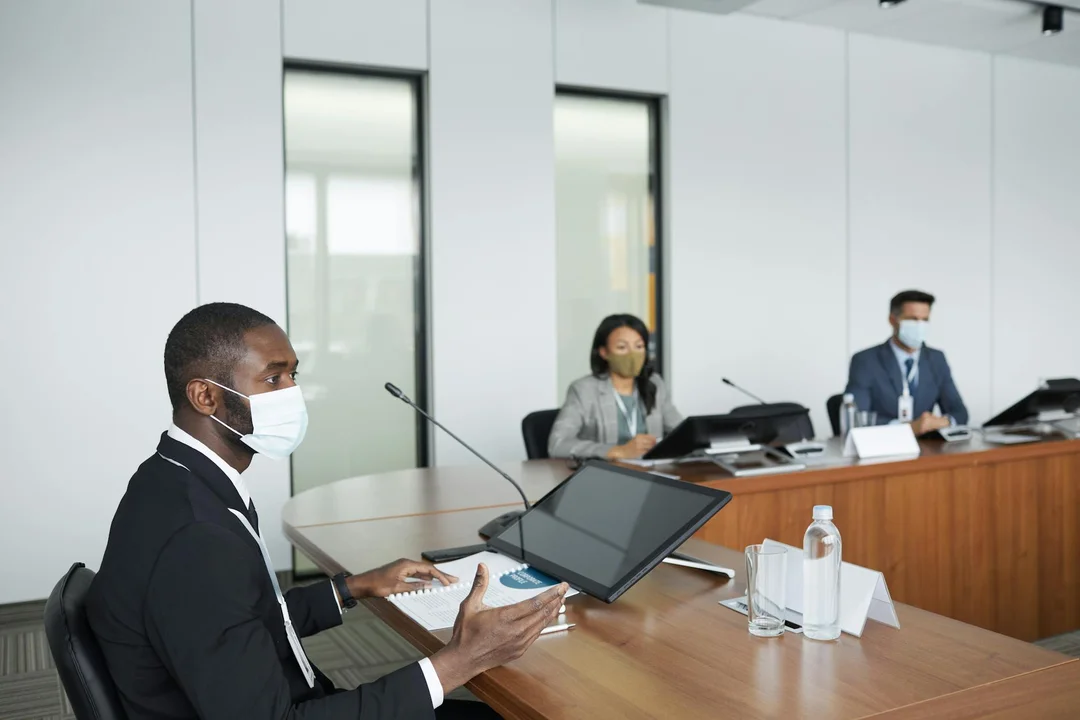 A diverse HOA board meeting in a modern conference room with participants wearing masks; a speaker at the foreground uses a laptop while others listen and take notes.