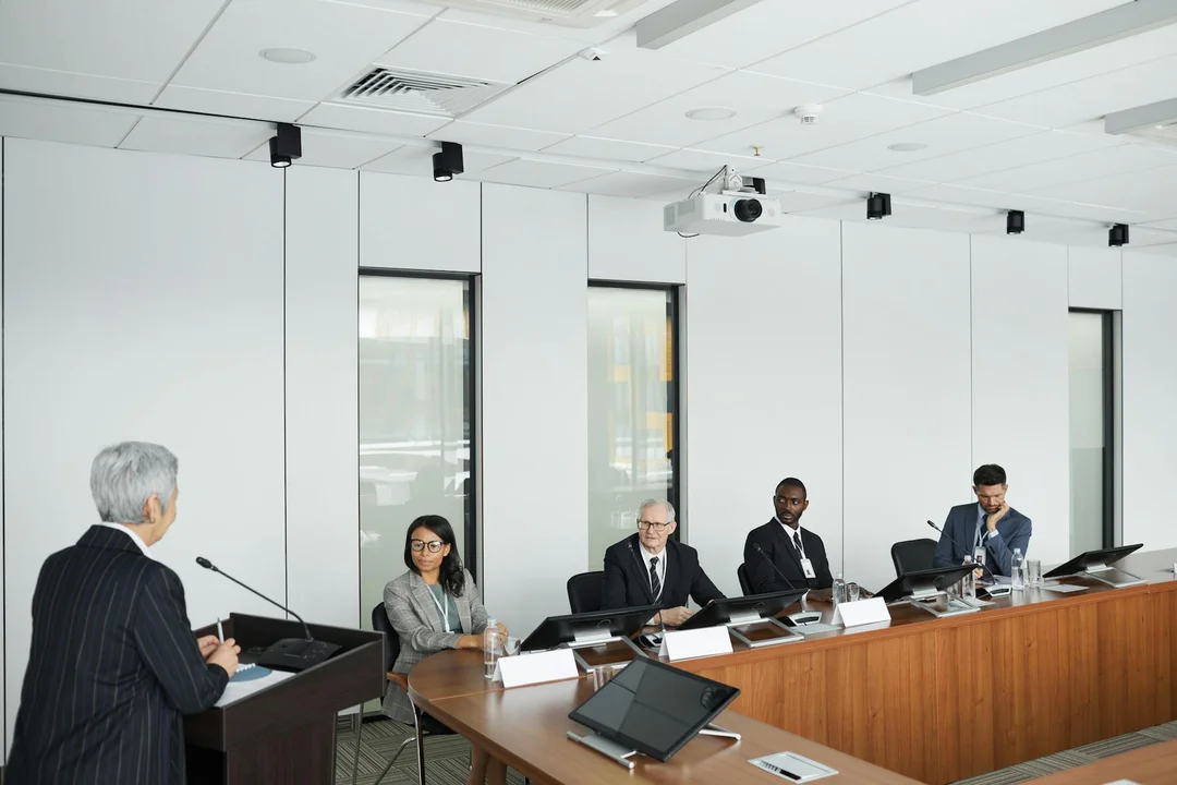 Group of HOA board members seated around a rectangular conference table in a bright, modern room; one person stands at a podium speaking to the group.