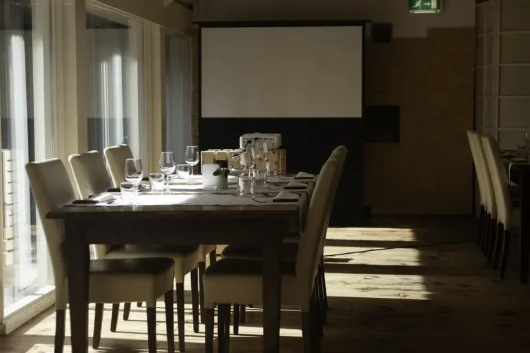 A sunlit conference room with a long wooden table set for a formal meeting, including wine glasses, napkins, and chairs facing a screen