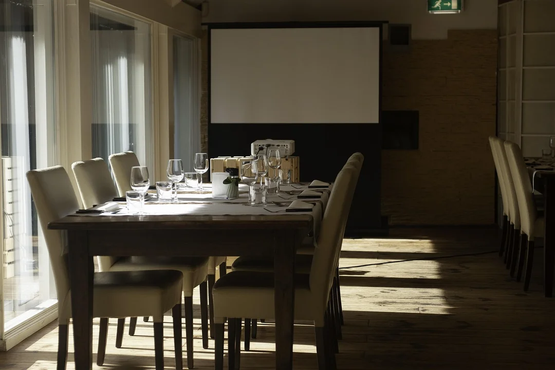 A sunlit conference room with a long wooden table set for a formal meeting, including wine glasses, napkins, and chairs facing a screen