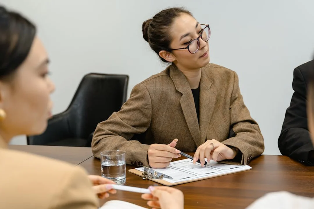 A woman wearing glasses and a brown blazer sits at a conference table, taking notes during an HOA board meeting while other members are present.