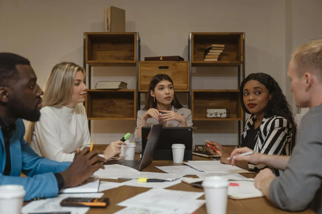 Diverse HOA board members sit around a conference table in a meeting room with laptops, notebooks, and coffee cups, discussing costs and benefits.