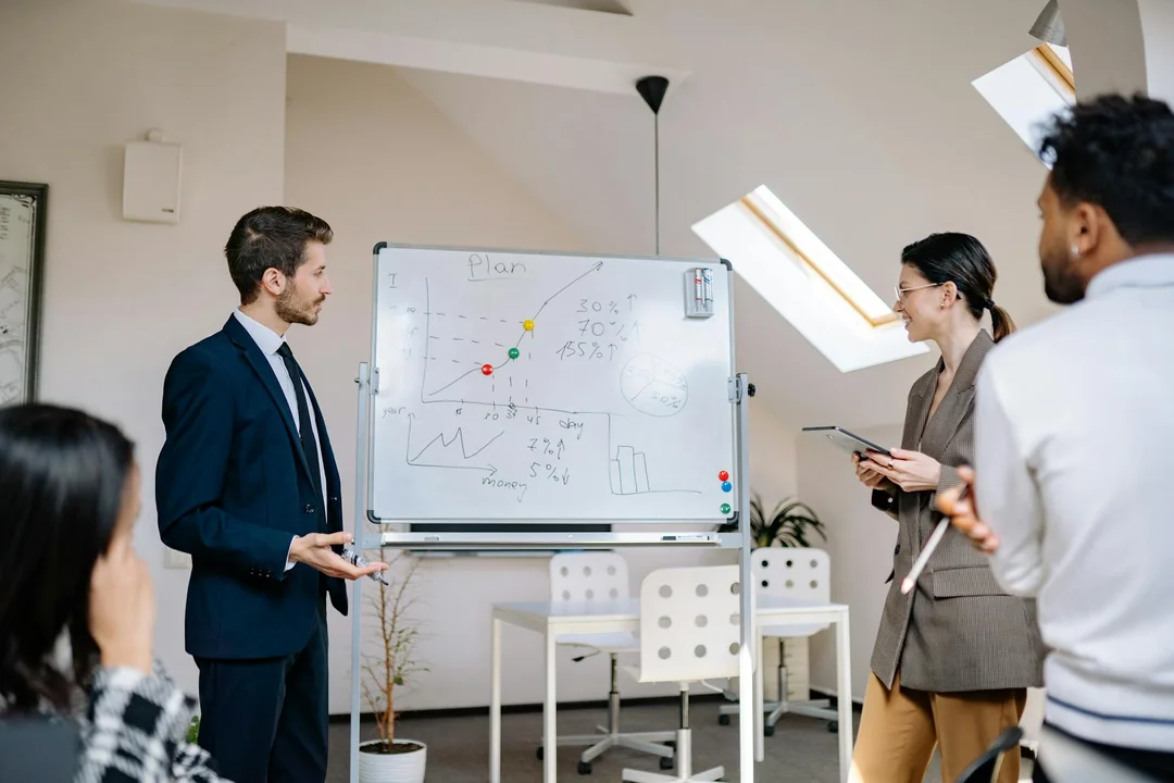 A group of professionals in a bright office discuss financial charts on a whiteboard during a meeting about HOA board compensation.