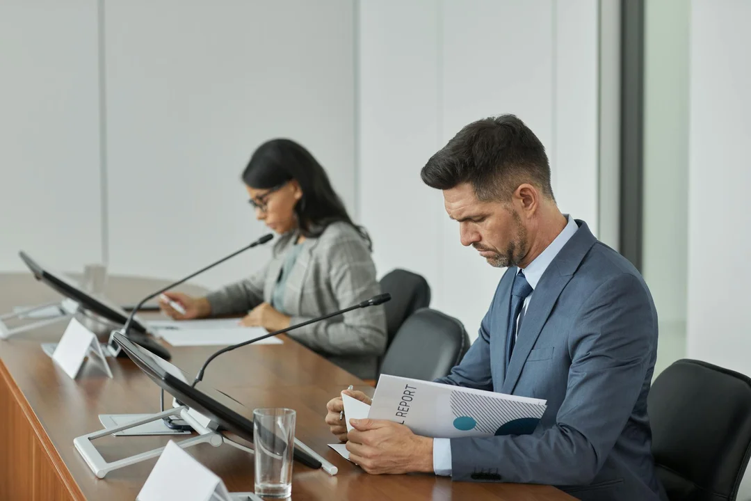 Two professionals sit at a long conference table reviewing documents during an HOA board meeting, with microphones and folders visible.