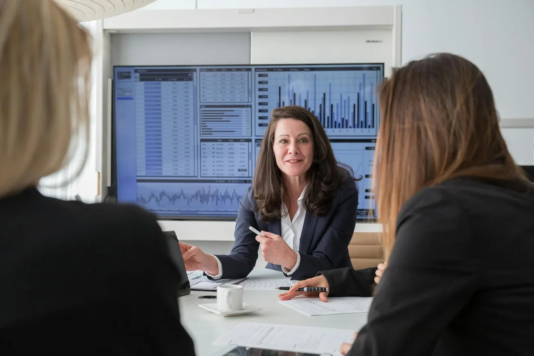 Professional woman leads HOA board meeting with two colleagues listening, while a monitor displaying charts and data is visible in the background.