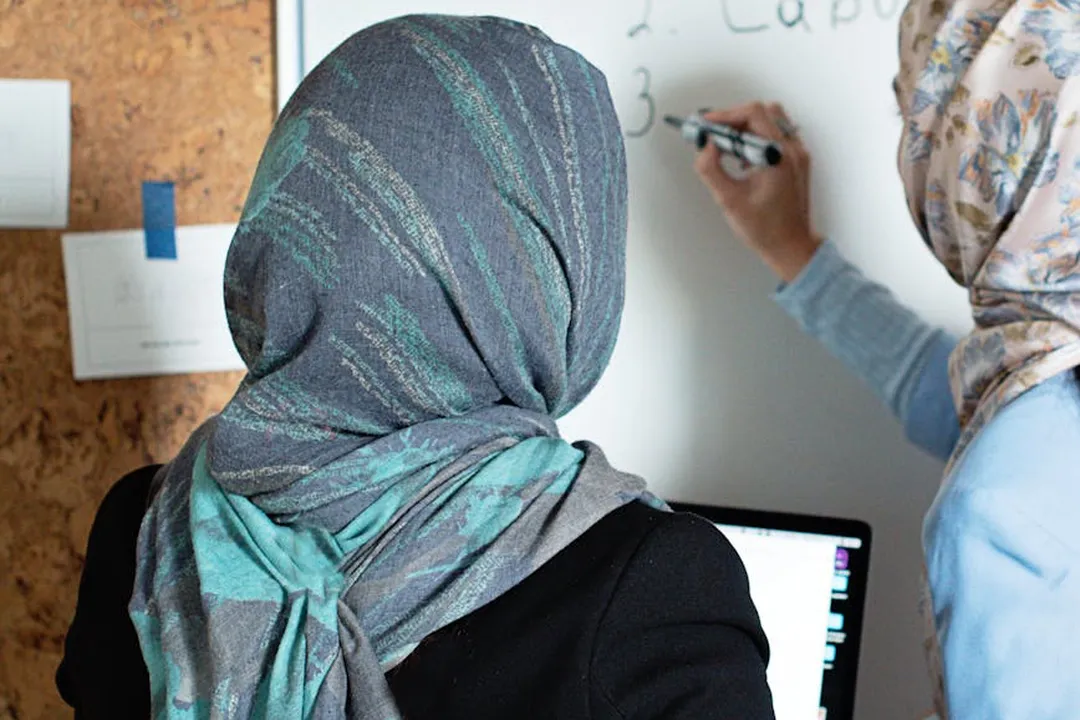 Two women wearing hijabs standing in front of a whiteboard and a laptop, collaborating on notes.