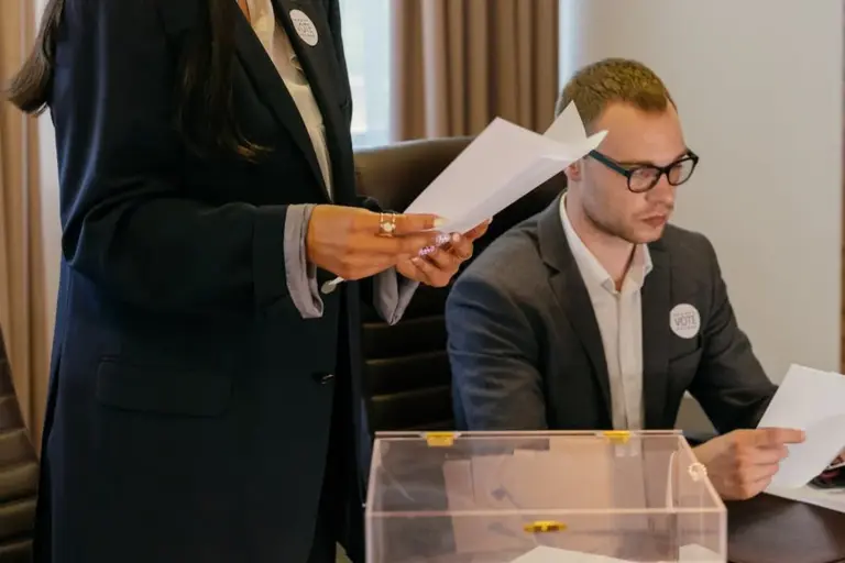 Two professionals in business attire at a meeting; a standing person holds documents while a seated person reviews papers, with a transparent ballot box on the table.