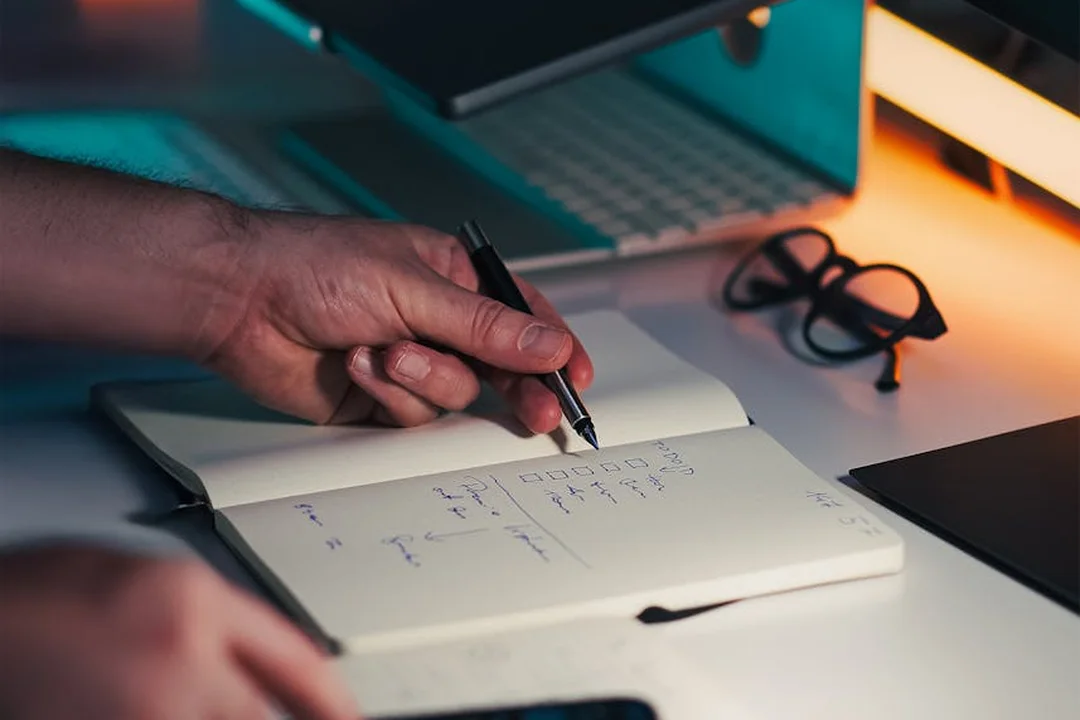 Close-up of a hand writing in a notebook on a desk with a laptop and glasses in the background.
