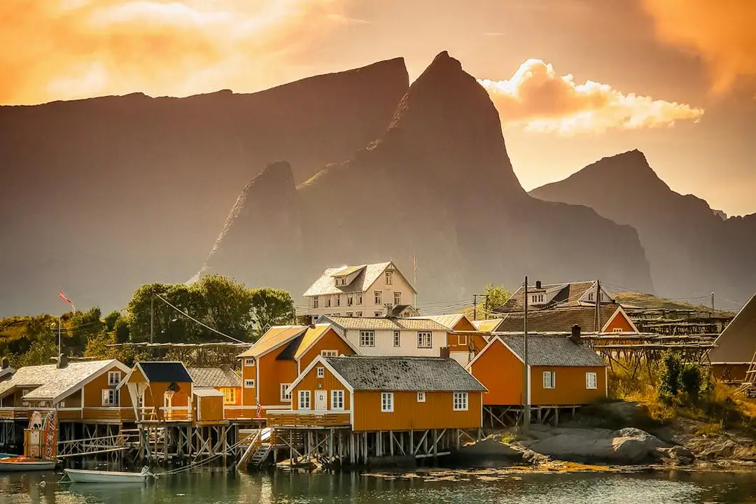 Sunset over a coastal village with colorful houses on stilts along the water, backed by mountains.
