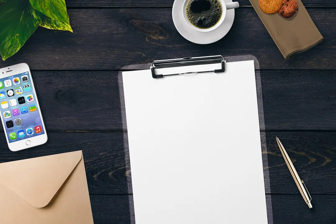 Clipboard with a blank sheet on a dark wooden desk, surrounded by a coffee cup, cookies, a smartphone, an envelope, a plant, and a pen.
