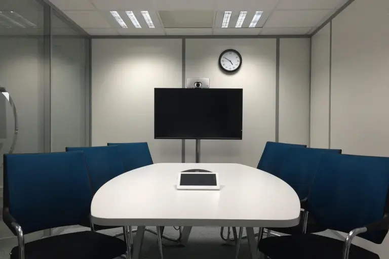 Modern HOA committee meeting room with an oval white table, blue chairs, a wall-mounted screen, and a clock on the wall.