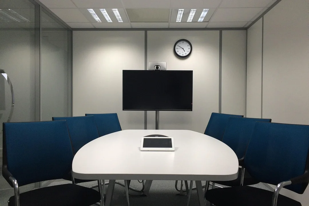 Modern HOA committee meeting room with an oval white table, blue chairs, a wall-mounted screen, and a clock on the wall.