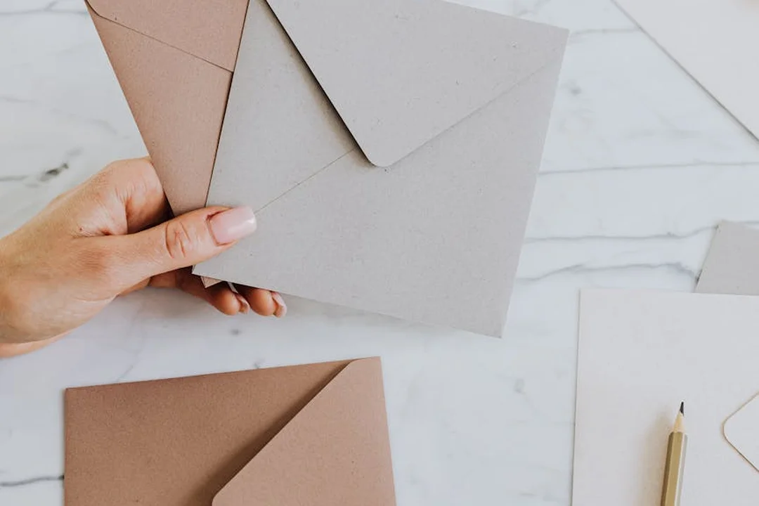 A hand holds brown and gray envelopes on a light marble desk, with additional stationery such as a pencil and papers nearby.